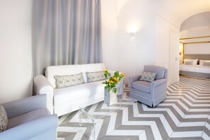 Moderni hotellihuone, jossa on valkoinen sohva, harmaa nojatuoli, lasinen sohvapöytä ja sängyssä pääty.