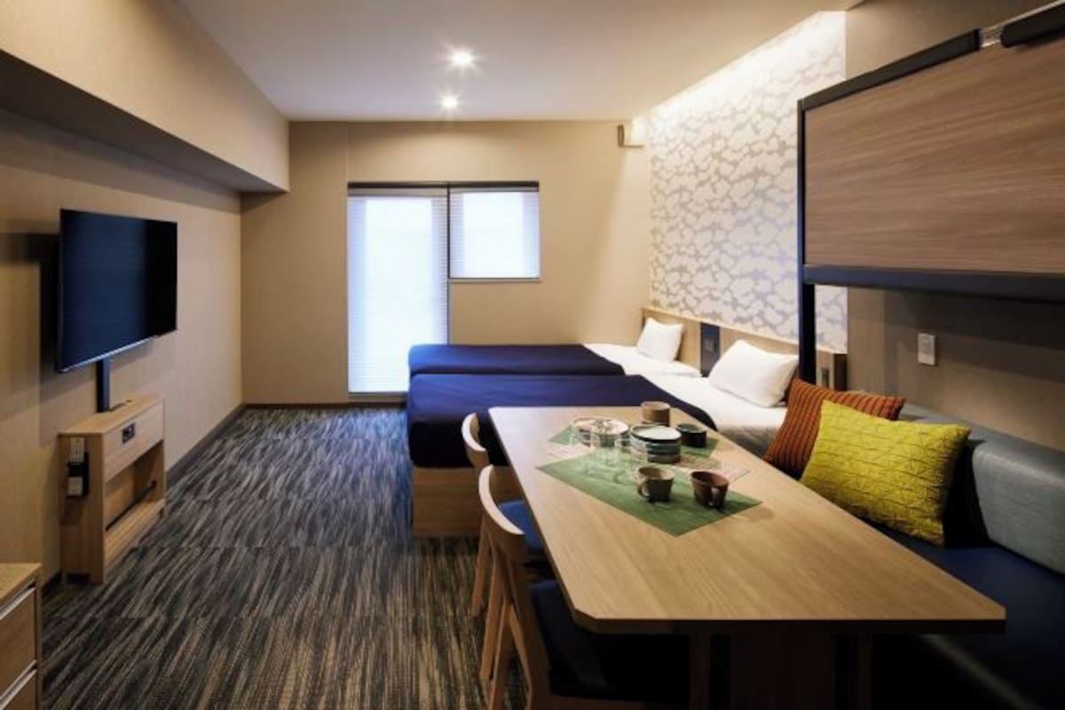 A modern hotel room with a bed, a dining table with chairs, a TV, and a window.