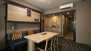 A compact hotel room with a kitchenette, a sofa with patterned cushions, a small dining table, and a refrigerator.