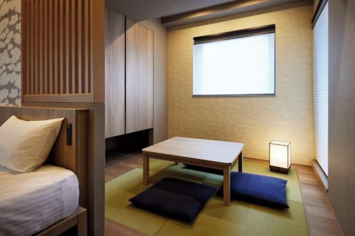 A compact room with a bed, a small table, a window with blinds, and a floor lamp.