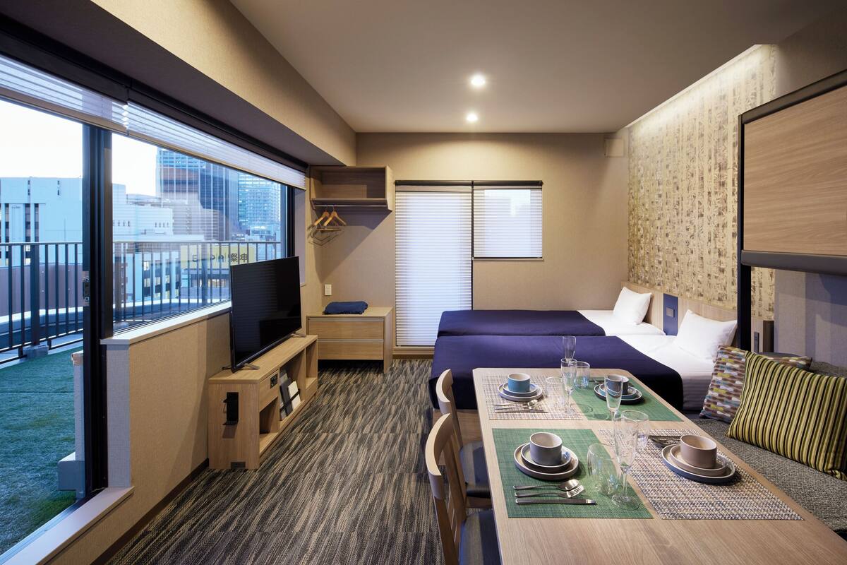 Ein Hotelzimmer mit Bett, Fernseher, Esstisch und Balkon mit Blick auf Gebäude.