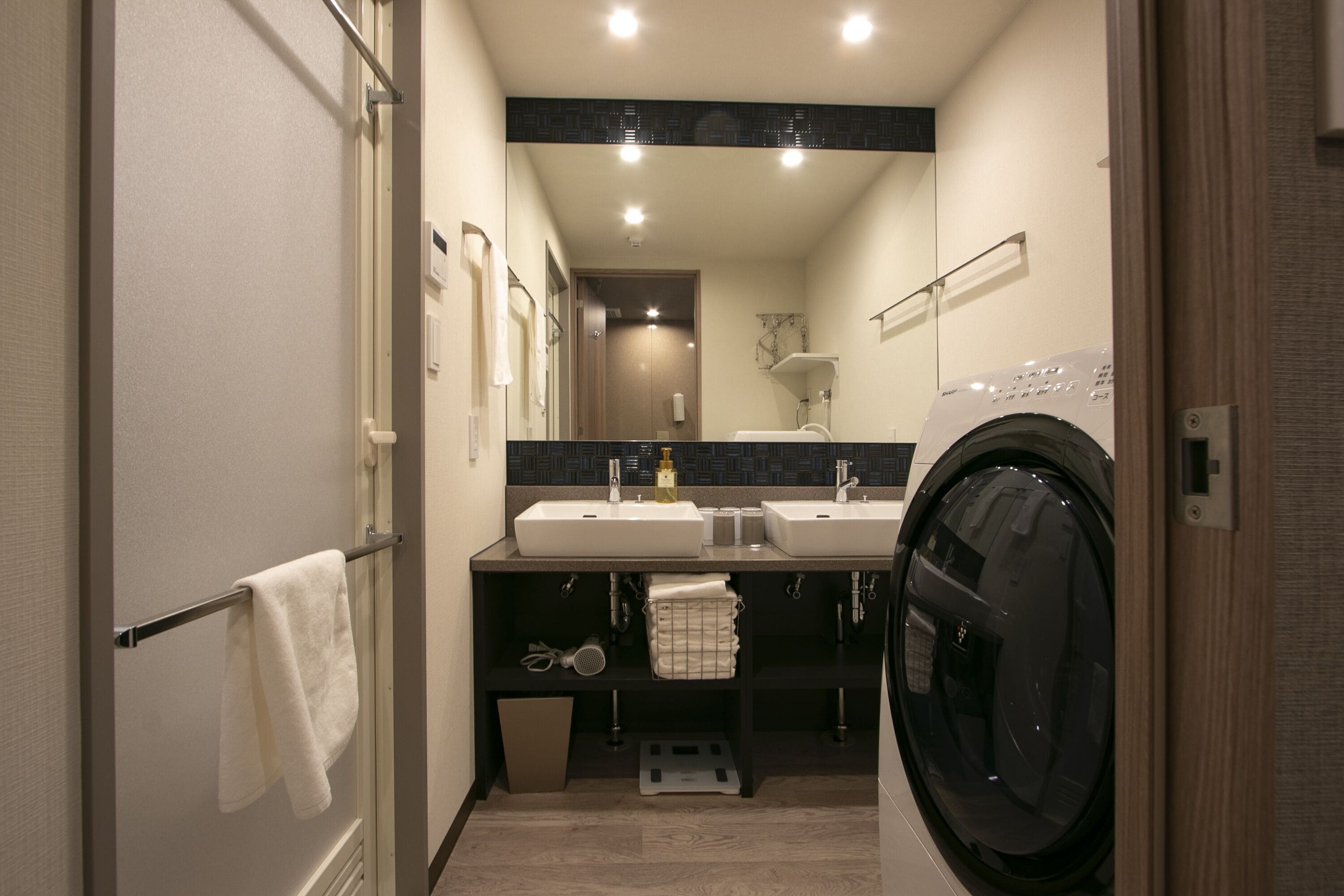 A compact hotel bathroom with two sinks, a washing machine, and a mirror.