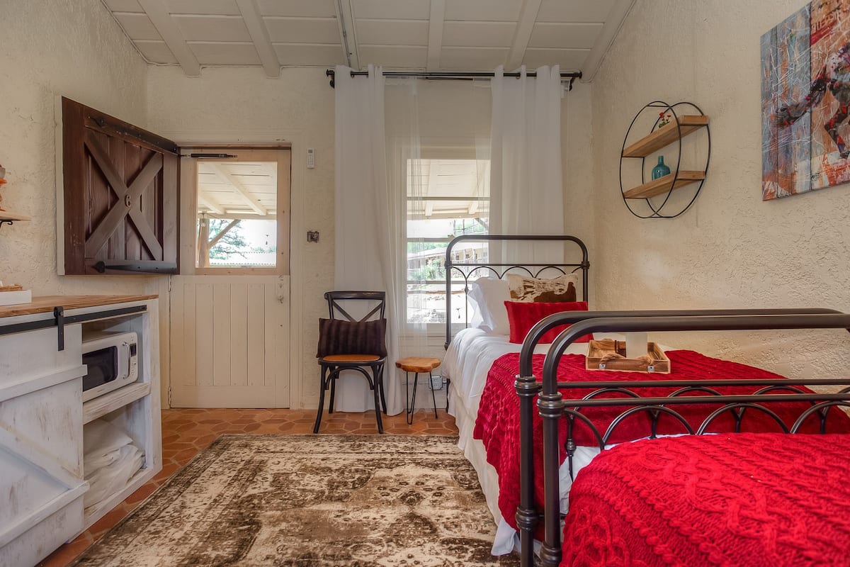 Un dormitorio con estructura de cama de metal, colcha roja, una silla de madera, un armario blanco y una ventana con cortinas.