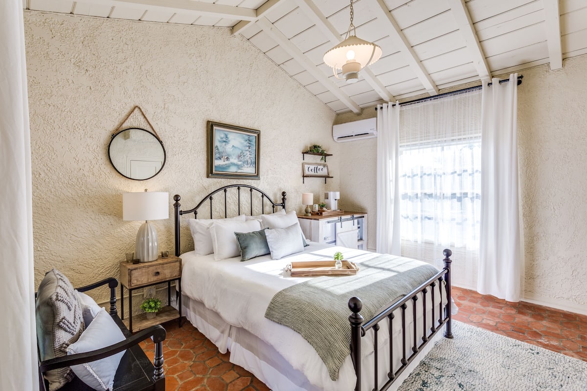 Un dormitorio con cama, mesita de noche, lámpara y un cuadro en la pared.