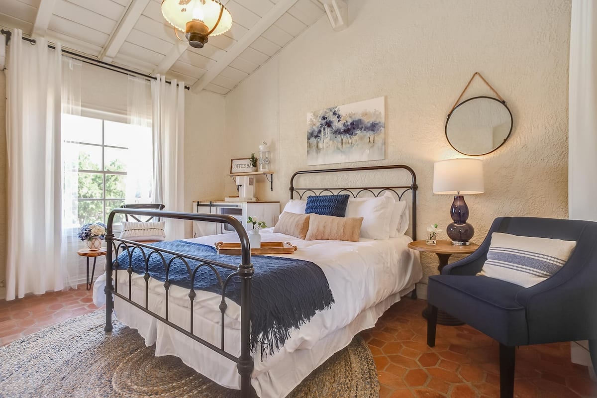 Un dormitorio con cama, una silla, una mesita de noche, una lámpara, un cuadro y una ventana con cortinas.