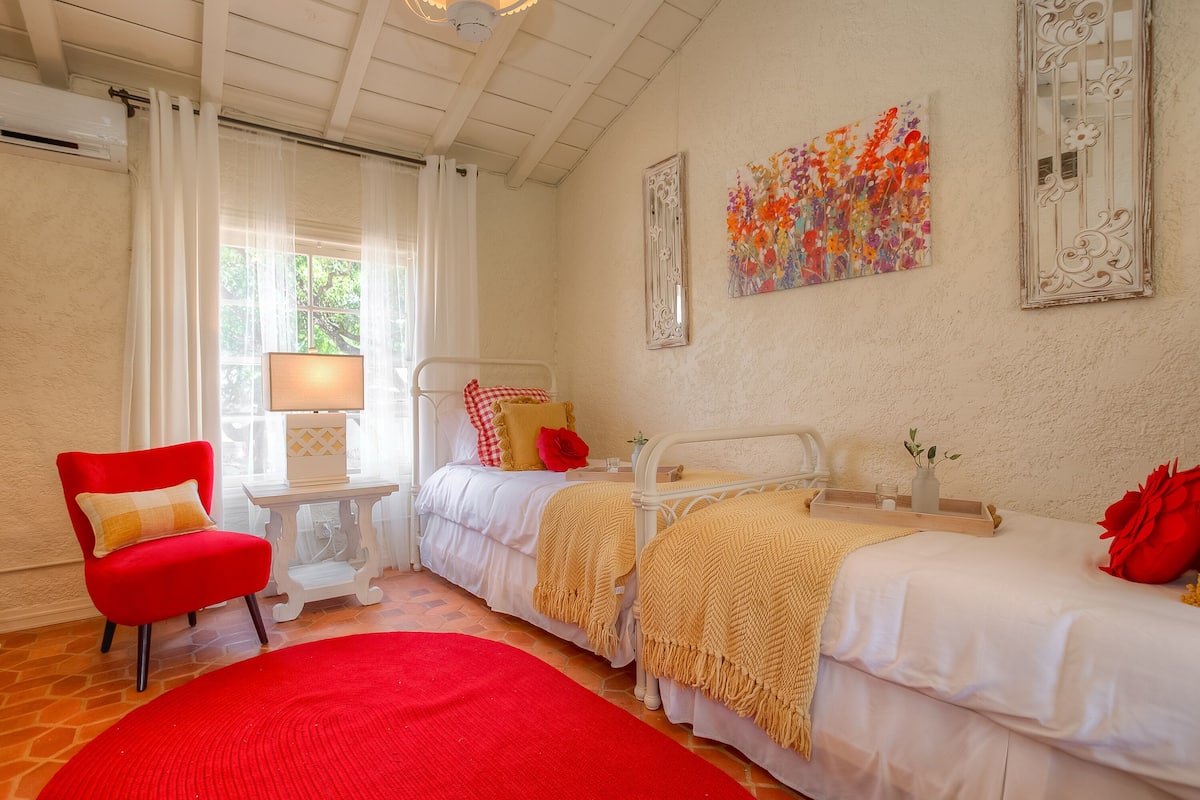 Un dormitorio con dos camas, una silla roja, una mesita de noche con lámpara y una ventana con cortinas.