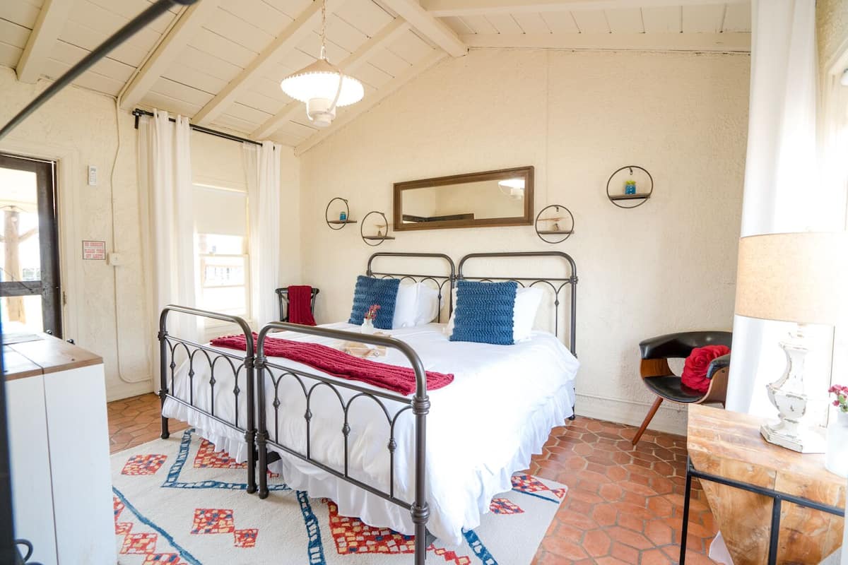 Un dormitorio con estructura de cama de metal, ropa de cama blanca, alfombra estampada, una silla de madera, una mesita con lámpara y un espejo en la pared.