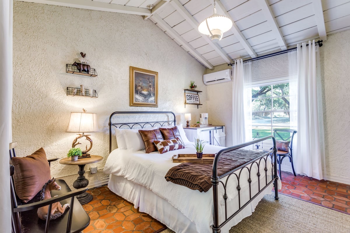 Un dormitorio con cama, mesitas de noche, lámpara, una silla y una ventana con cortinas.