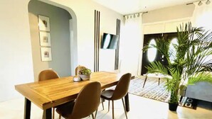 A dining area with a wooden table and four chairs, a TV mounted on the wall, and a large potted plant.