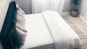 A neatly made bed with white bedding and a quilted blanket, a dark headboard, sheer curtains, a woven basket, and a potted plant.