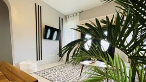 A modern living room with a wooden table, a patterned rug, a large potted plant, and a flat-screen TV mounted on the wall.