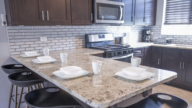 A kitchen with a granite countertop, dark cabinets, a microwave, a stove, and bar stools.