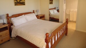 A neatly made bed in a hotel room with a wooden headboard, a bedside table with a lamp, a nightstand, and a mirror reflecting the room.