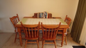 A dining table with a placemat, surrounded by four wooden chairs. A bookshelf with books is against the wall.