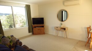 A living room with a sofa, a TV on a wooden stand, a small desk with a mirror, and a window with curtains.