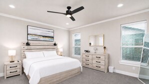 A bedroom with a bed, bedside tables, a dresser, a ceiling fan, a large window with blinds, and a wall-mounted artwork.