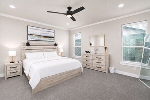 Une chambre à coucher comprenant un lit, des tables de chevet, une commode, un ventilateur de plafond, une grande fenêtre avec des stores et une œuvre d’art accrochée au mur.