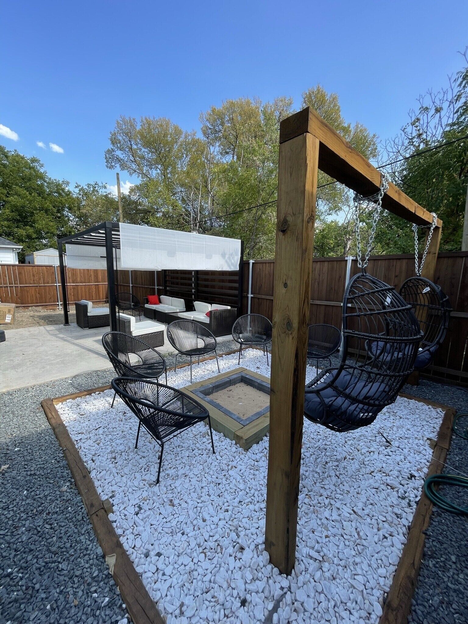 Un jardin avec une pergola en bois, un fauteuil suspendu et un espace salon couvert, équipé de tables et de chaises.
