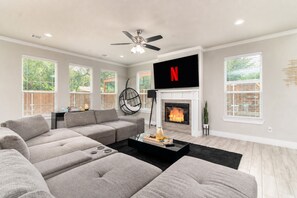 Un salon moderne avec un canapé d’angle, une cheminée, un fauteuil suspendu et un téléviseur à écran plat fixé au-dessus de la cheminée.