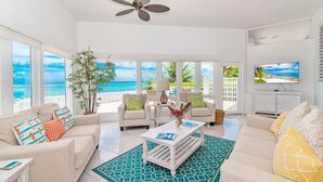 A spacious living room with a large window overlooking the ocean, a ceiling fan, and a television on the wall.