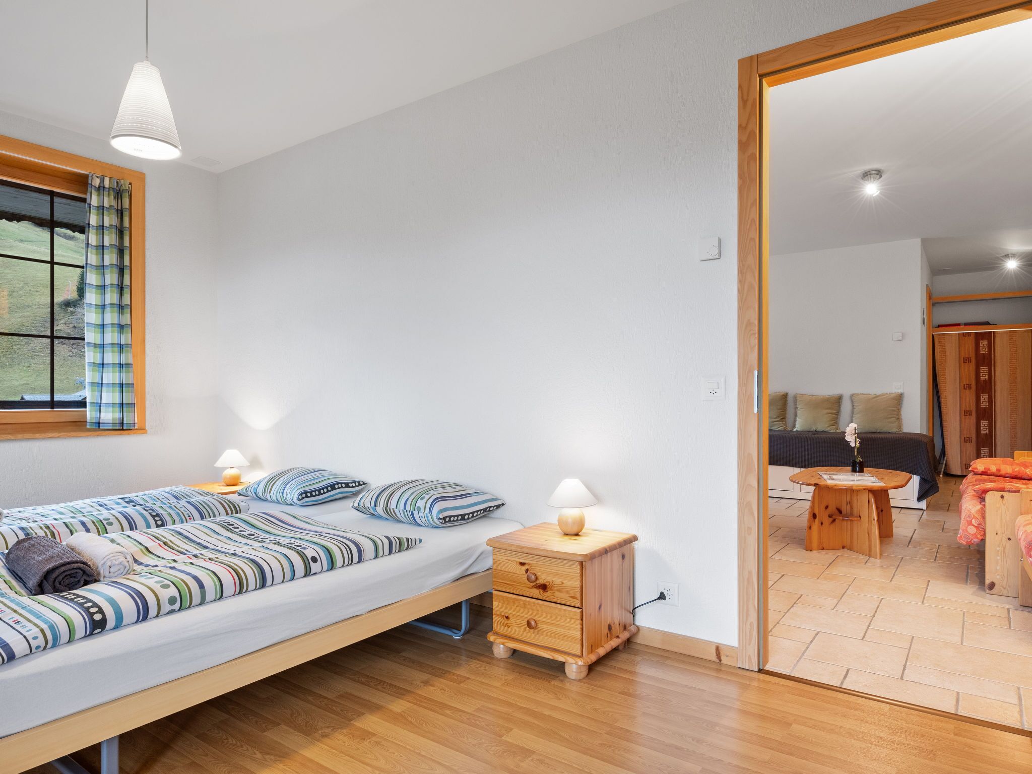 A modern bedroom with a single bed, bedside tables, a wooden nightstand, and a view through an open door to another room.