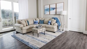 Een woonkamer met een beige modulaire bank, een gestreept voetenbankje, een blauw-wit vloerkleed, een vloerlamp en een verzameling ingelijste kunstwerken aan de muur.