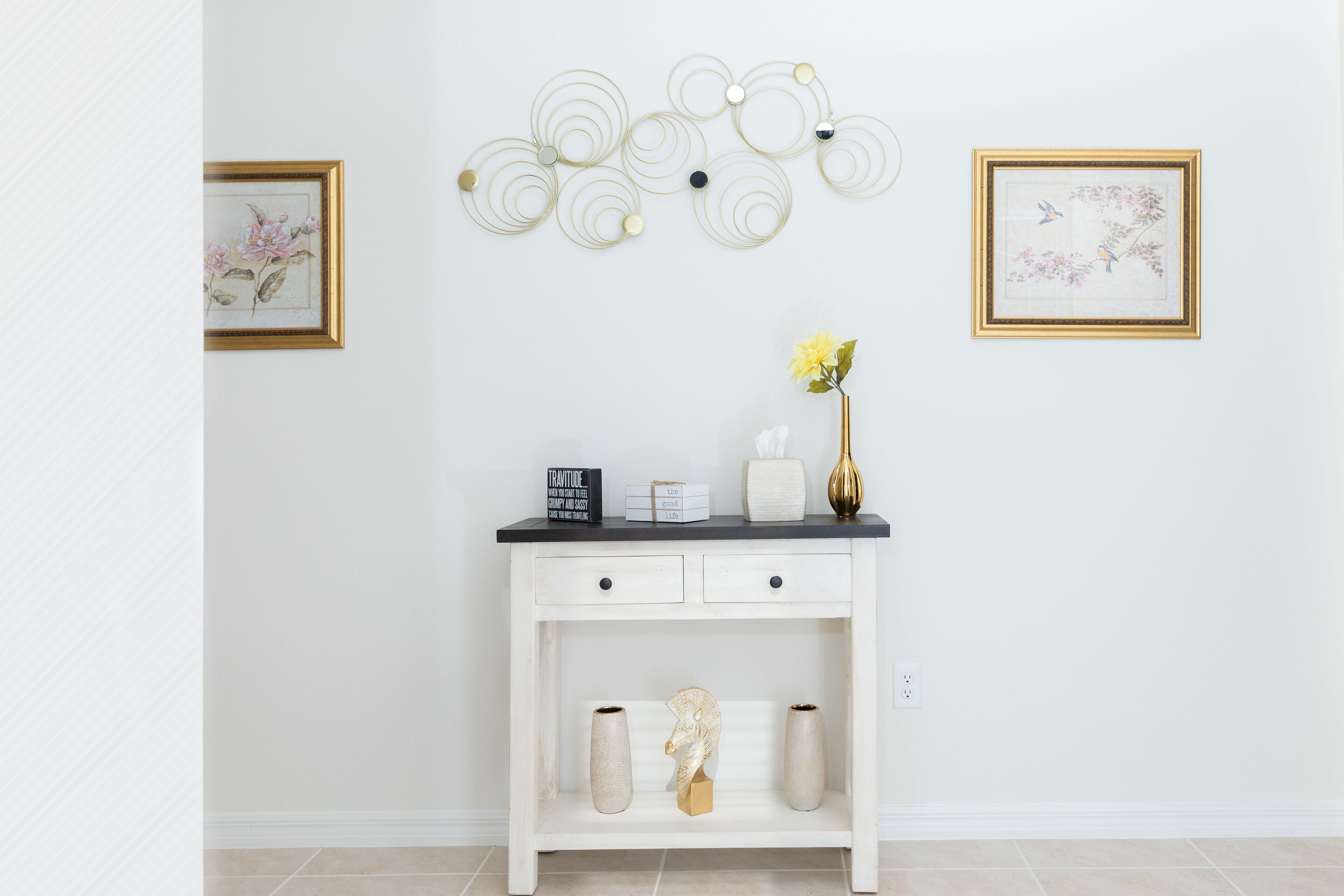 A white console table with a black top, a vase with yellow flowers, and a decorative wall piece above it.