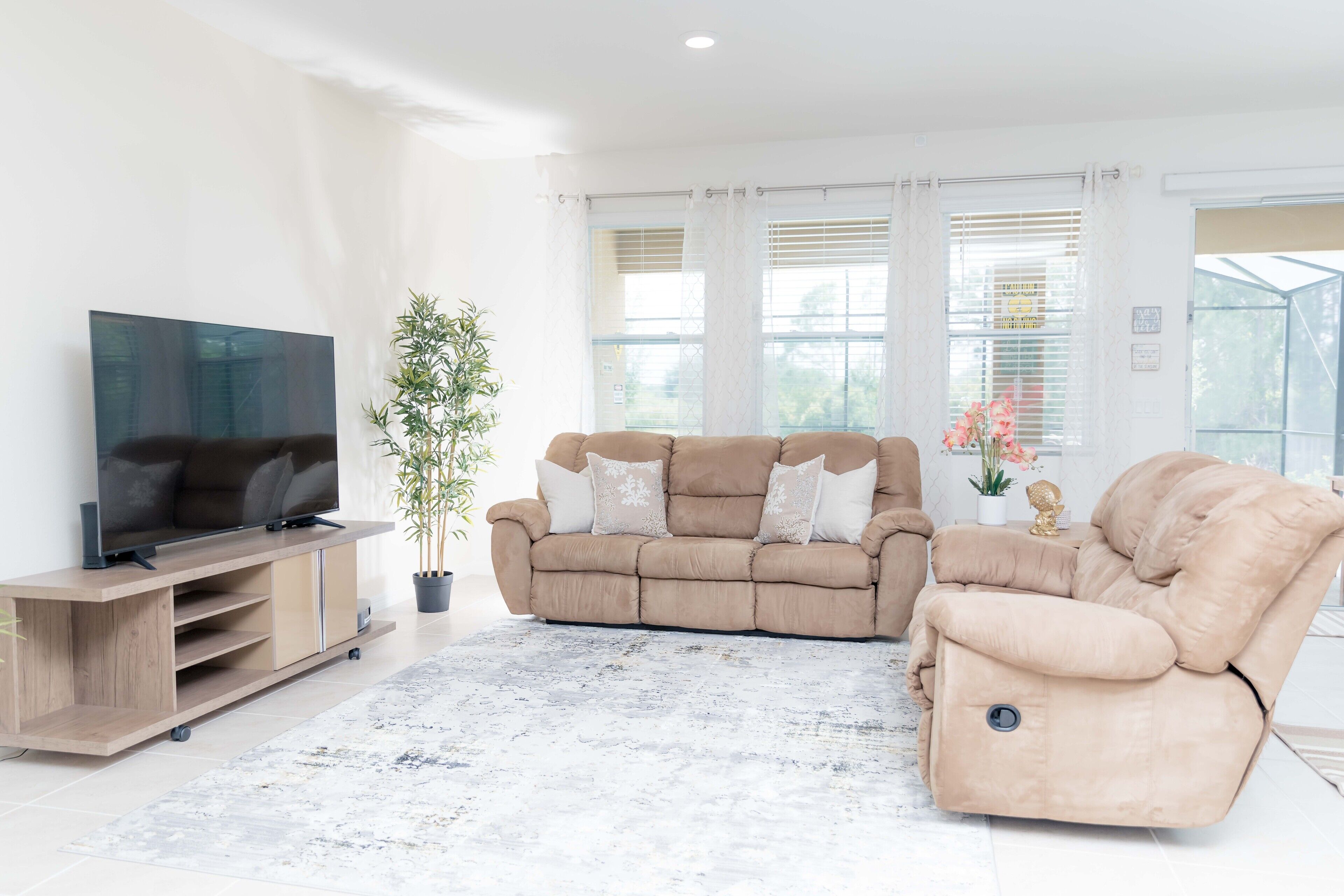 A modern living room with a beige sofa, a TV on a wooden stand, a potted plant, and a view through large windows.