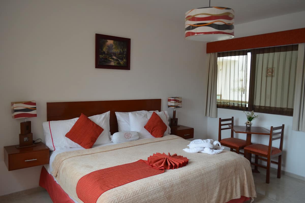 A neatly arranged hotel room with a large bed, a bedside table with a lamp, a small table with chairs, and a window with curtains.