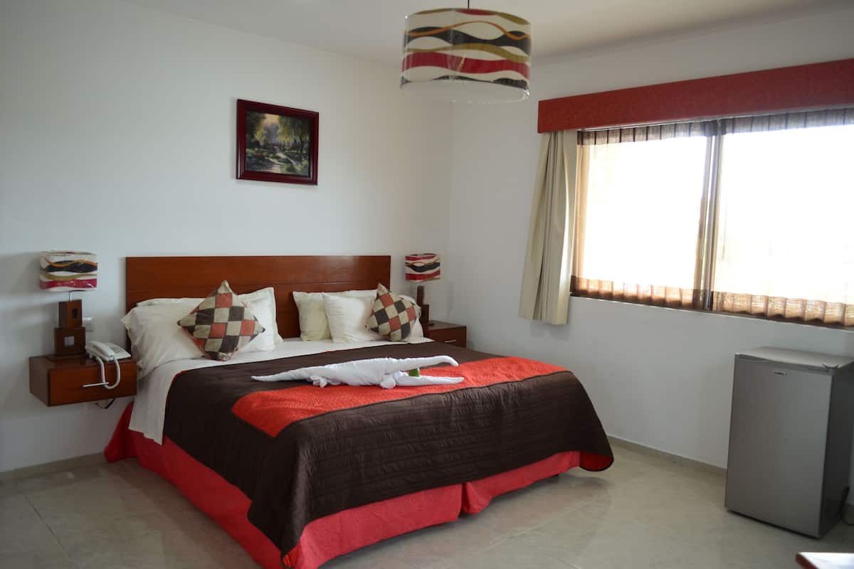 A neatly arranged bedroom with a large bed, a bedside table with a lamp, a small refrigerator, and a window with curtains.