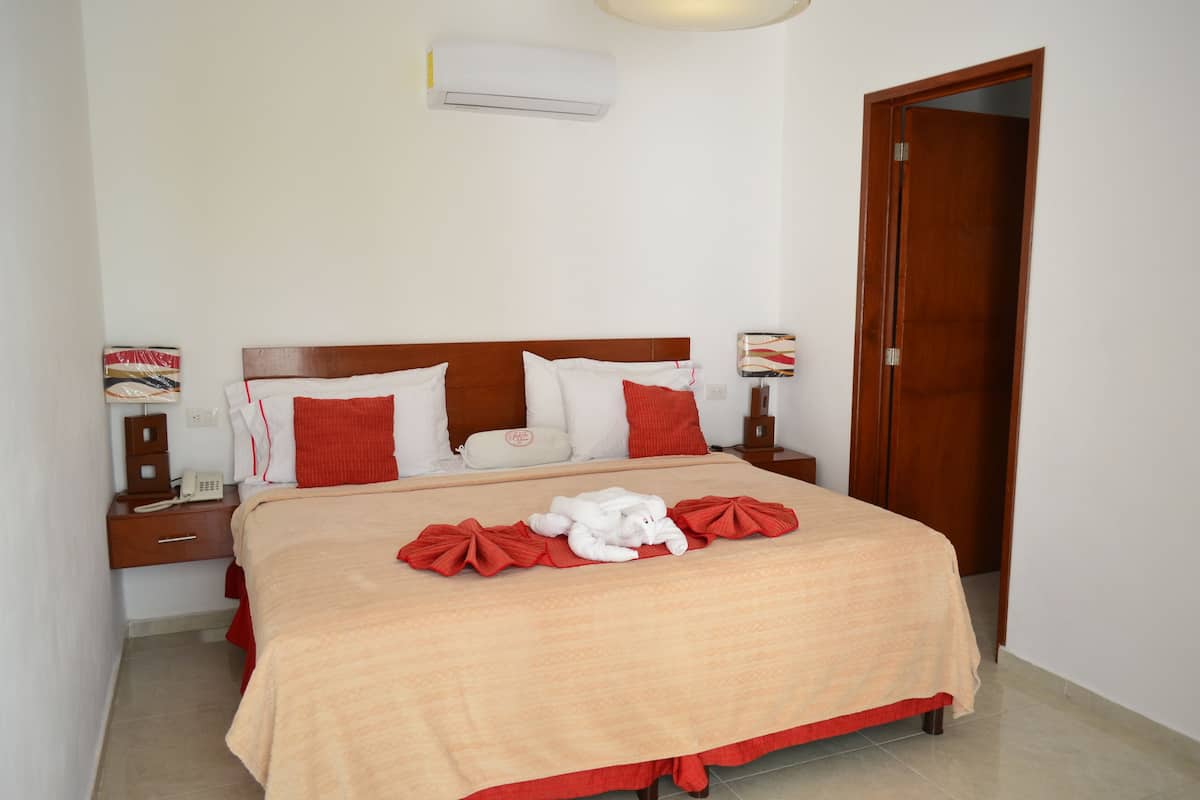 A neatly made bed with a beige bedspread, red pillows, and a white towel. There is a wooden nightstand with a lamp and a telephone.