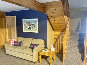 Un salon avec un canapé beige, deux coussins ornés du mot « HOME », une carte encadrée des États-Unis et un escalier en bois.