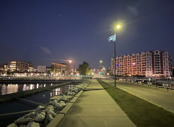 A waterfront promenade with a canal, buildings, and a flagpole. - Marina Life II - 28 Carver @ Reef Point Marina (Racine)