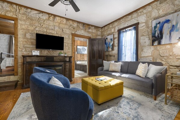 Un salon avec des murs en pierre, une cheminée, un canapé, un pouf jaune et un téléviseur à écran plat.