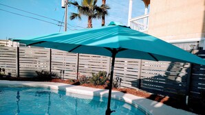 En swimmingpool med en blå parasol, omgivet af et træhegn og en palme.