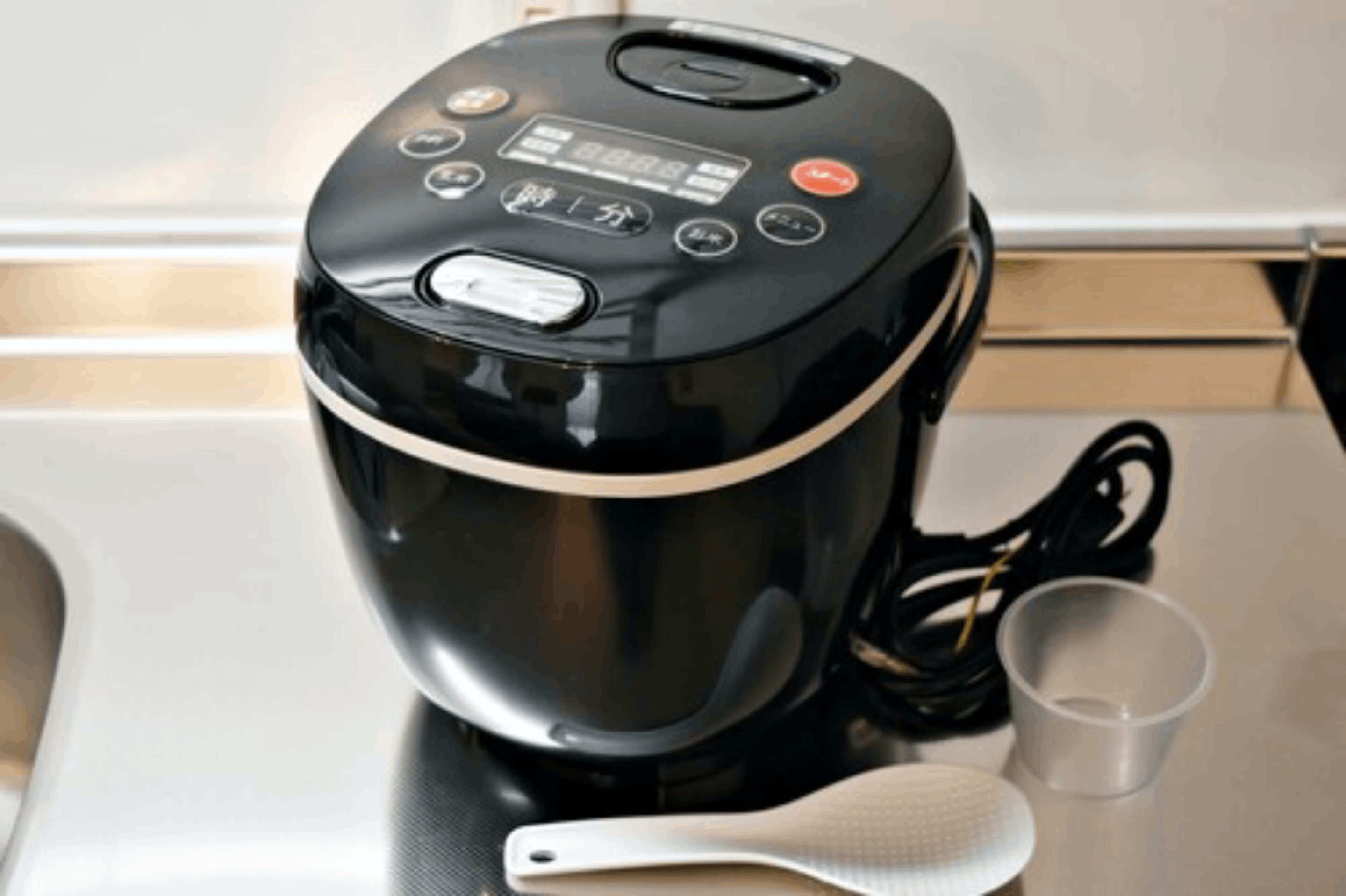Una tetera eléctrica negra con pantalla digital, un cable de alimentación y una cuchara de silicona blanca sobre una encimera de cocina.