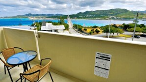 A balcony with wicker chairs and a small table, overlooking a coastal view with mountains and a clear sky. - Ocean Terrace Sesoko 101 - Vacation Stay 28609V (Motobu)