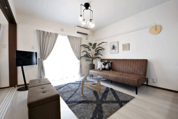 A modern living room with a sofa, a glass coffee table, a TV, and a large window with curtains. - Ocean Terrace Sesoko 101 - Vacation Stay 28609V (Motobu)