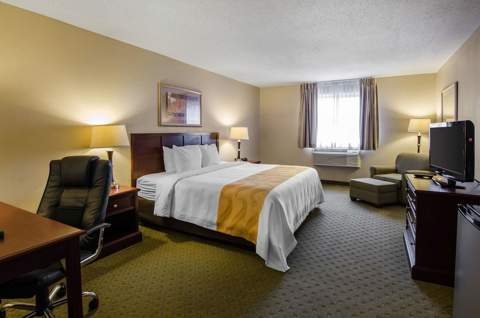 Quarto de hotel com cama, escrivaninha, cadeira, televisão e janela com cortinas.