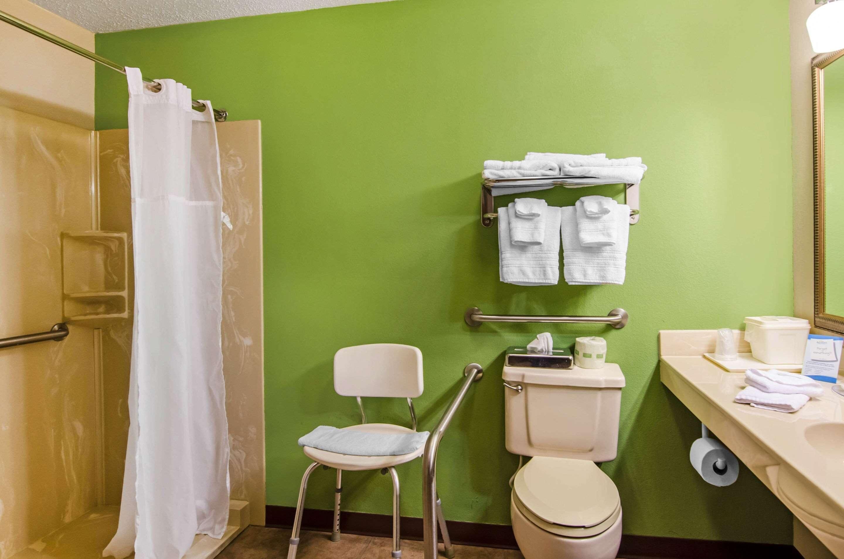 A bathroom with a green wall, a white toilet, a shower with a white curtain, a white chair, and a towel rack with folded towels.