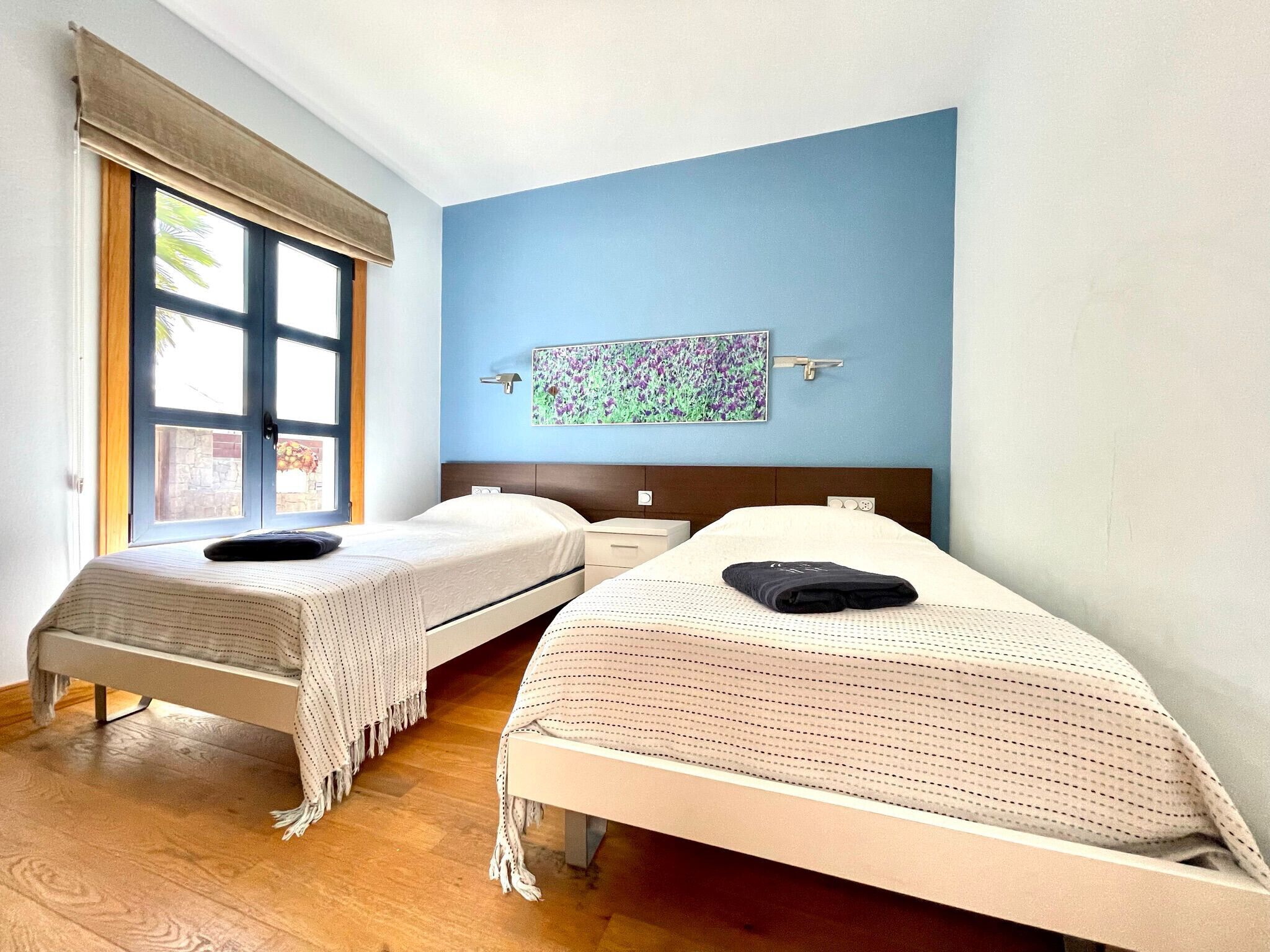 A room with two single beds, a window with a view, a wall-mounted artwork, and a wooden floor.