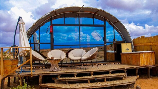 Une grande structure arquée en verre, dotée d’une terrasse en bois, de deux fauteuils en osier et d’une petite table.