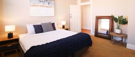 A bedroom with a large bed, bedside tables with lamps, a painting on the wall, a door, and a potted plant.