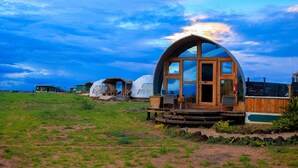 Eine Holzhütte mit einer großen Glasfront, umgeben von Jurten in einem grasbewachsen Feld.