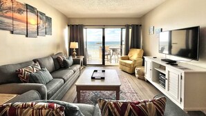 A hotel room with a sofa, armchair, TV, and a view of the sea through the balcony doors. - Tropical Winds 303 2 Bedroom Condo by RedAwning (Gulf Shores)