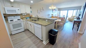 A modern kitchen with white cabinets, a granite countertop, and a dishwasher. There is a dining area with a table and chairs, and a trash can. - Tropical Winds 303 2 Bedroom Condo by RedAwning (Gulf Shores)