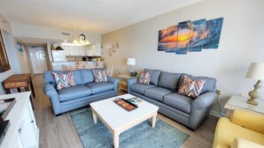 A living room with a sofa set, a coffee table, and a dining area with a kitchen in the background. - Tropical Winds 303 2 Bedroom Condo by RedAwning (Gulf Shores)