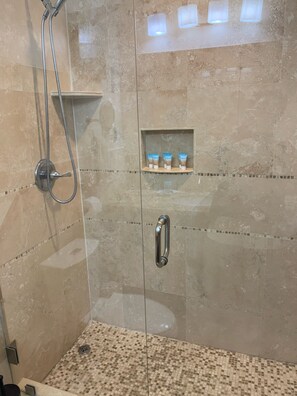 Zona de ducha con puerta de vidrio, ducha de metal y repisa con artículos de aseo.