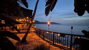 Eine Terrasse direkt am Strand mit Sitzgelegenheiten im Freien, einer Brüstung und Blick auf das Meer und vorgelagerte Inseln.
