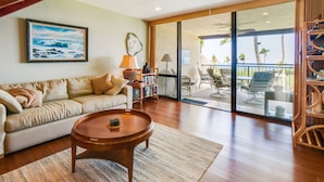 A living room with a sofa, coffee table, and bookshelf. There is a sliding glass door leading to an outdoor area with chairs and a view of palm trees. - 2 Story top level AC unit 2 bdrms w/Large Loft with 2 twin beds & bathrm SUNSETS (Kailua-Kona)